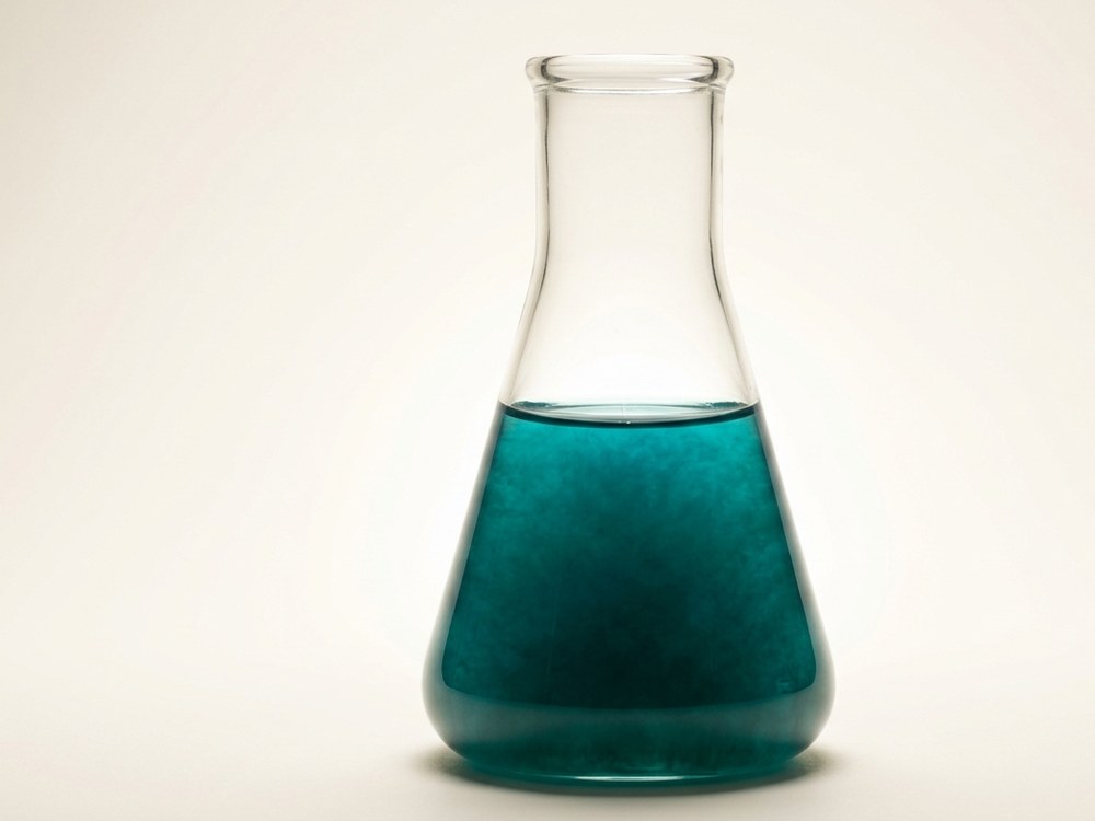 A laboratory flask holding a dense blue-green microalgae culture, backlit to show the depth of the suspension.