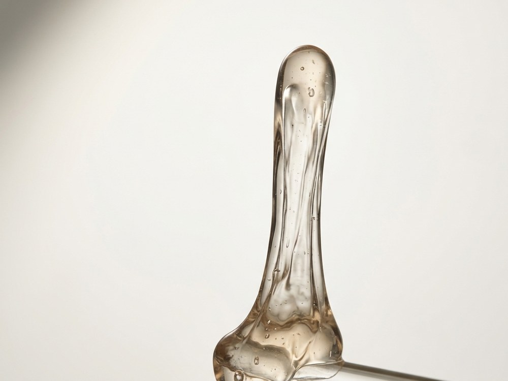 A translucent amber gel drawn upward into a thick thread, showing high viscosity and elasticity against a neutral background.